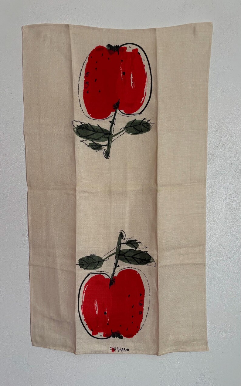 May include: A rectangular beige tea towel featuring two vertically aligned red apple illustrations with black outlines and dark green leaves. The apples are rendered in a painterly style, and the text "USVA" is printed in small black letters at the bottom.