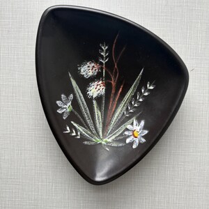 May include: A black, triangular-shaped ceramic dish with a floral design. The hand-painted design features white and yellow flowers, green leaves, and brown stems. The dish has a matte finish and is likely vintage.