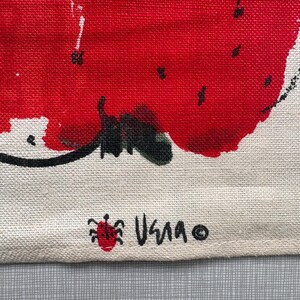 May include: A textured off-white fabric featuring a large, vibrant red painted strawberry graphic with black seeds and a green stem. The bottom corner displays a small red ladybug icon next to the black handwritten text VERA and a copyright symbol.