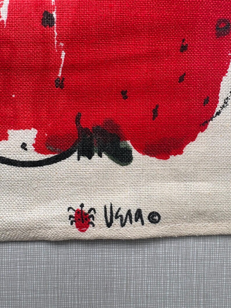 May include: A textured off-white fabric featuring a large, vibrant red painted strawberry graphic with black seeds and a green stem. The bottom corner displays a small red ladybug icon next to the black handwritten text VERA and a copyright symbol.