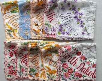 May include: A collection of ten white fabric handkerchiefs with scalloped edges, each featuring a state name in cursive script, a map outline, and a floral illustration. The states include Alabama, Alaska, California, Illinois, and North Dakota.
