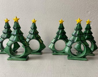 May include: Six green ceramic Christmas tree napkin rings with yellow star toppers. Each tree has a circular opening in the center. The napkin rings are arranged on a light-colored surface.