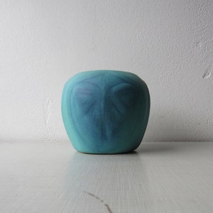 Puede incluir: Un jarrón de cerámica de color turquesa, de forma redondeada y superficie texturizada. El jarrón presenta un sutil patrón azul más oscuro en la parte delantera, que se asemeja a una cara estilizada. El jarrón se exhibe sobre una superficie blanca contra una pared blanca.