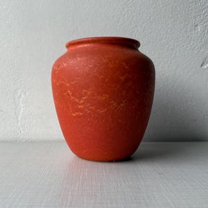 Puede incluir: Un jarrón de color terracota con una superficie texturizada y una forma redondeada. El jarrón tiene un cuerpo ancho que se estrecha hacia una base más pequeña y un borde ligeramente ensanchado. El color es un rojo-naranja vibrante con sutiles vetas amarillo-naranja.