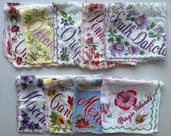 May include: A collection of white fabric handkerchiefs with scalloped edges, each featuring colorful floral illustrations and printed state or regional names including Oklahoma, Oregon, Pennsylvania, South Dakota, Wisconsin, Canada, Mexico, and Virgin Islands.