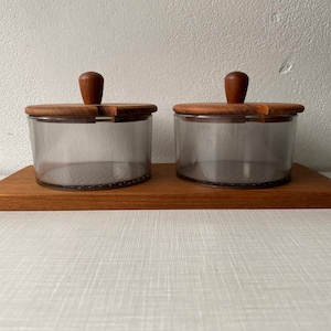 Puede incluir: Dos recipientes de vidrio transparente con tapas y pomos de madera sobre una bandeja rectangular de madera. Los recipientes tienen un tinte ahumado y están diseñados con una estética minimalista. Los elementos de madera tienen un tono cálido marrón rojizo.