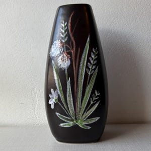 May include: A dark brown ceramic vase with a painted floral design. The vase features white flowers, green leaves, and brown stems. The vase is a tall, oval shape, and the floral design is centered on the front.