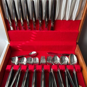 May include: A wooden box with a red velvet interior containing a set of silverware with black handles. The silverware includes forks, knives, and spoons. The box has a label that reads "18/10 Stainless Steel, Guaranteed Finish, Made in England, Towle"