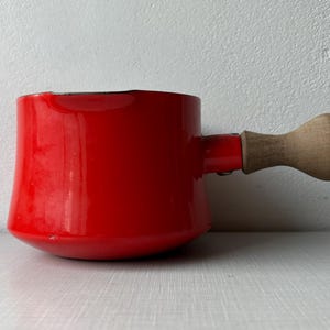 May include: A small, red enamel saucepan with a wooden handle. The pot has a rounded base and a cylindrical body. The handle is attached to the side of the pot and is made of light-coloured wood. The saucepan is sitting on a white surface.