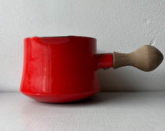 Op de afbeelding: Een kleine, rode emaille steelpan met een houten handvat. De pot heeft een afgeronde bodem en een cilindrisch lichaam. Het handvat is aan de zijkant van de pot bevestigd en is gemaakt van lichtgekleurd hout. De steelpan staat op een wit oppervlak.