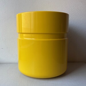 Puede incluir: Un macetero cilíndrico de color amarillo brillante con un acabado liso y brillante. El macetero tiene un borde sutil alrededor de la parte superior e inferior, creando un efecto de dos niveles.
