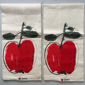 May include: Two rectangular off-white kitchen towels, each featuring a large, stylized red apple illustration with black ink outlines and dark green leaves. The bottom of each towel displays a small red apple icon and the text "USIA" in black.