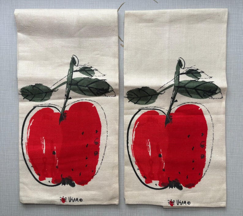 May include: Two rectangular off-white kitchen towels, each featuring a large, stylized red apple illustration with black ink outlines and dark green leaves. The bottom of each towel displays a small red apple icon and the text "USIA" in black.