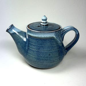 Könnte beinhalten: Eine handgefertigte Keramik-Teekanne in Blautönen. Die Teekanne hat einen abgerundeten Körper, einen gebogenen Ausguss und einen Henkel. Der Deckel ist mit einem kleinen, dekorativen Knopf versehen. Die Glasur hat ein strukturiertes Aussehen.