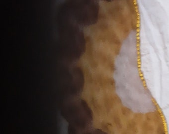 May include: A crescent-shaped item with a textured surface, featuring a gradient of colors from dark brown to light yellow. A thin, gold-colored trim runs along one edge, and the item rests on a white fabric.
