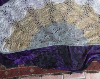 May include: A white, beige, and gray knitted shawl with a lacy pattern. The shawl is folded and draped over a purple fabric.