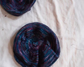 May include: Two knitted items, possibly hats or bowl cozies, in shades of blue, purple, and red. The items are circular with a textured pattern, resting on a white fabric surface. The yarn appears to be a variegated blend.