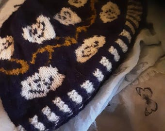 May include: A blue knitted hat with a white and gold pattern of ghosts. The hat is folded and lying on a light blue fabric with a butterfly pattern.