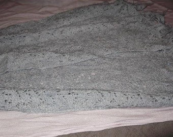 May include: A gray crocheted shawl with a delicate pattern. The shawl is folded and lying on a pink surface.