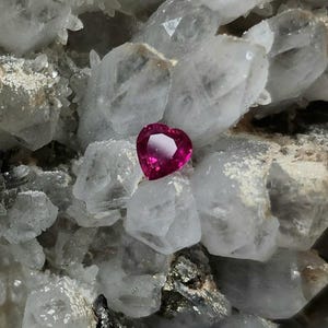 May include: A faceted heart-shaped gemstone with a vibrant deep pink color, featuring precise geometric cuts that reflect light, resting against a cluster of translucent, jagged, and crystalline white mineral formations.