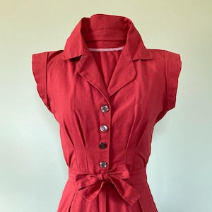 May include: A red romper with a collared neckline, short sleeves, and a button-down front. The romper features a waist tie and rolled-up shorts. The fabric appears to be a lightweight material, suitable for warm weather.