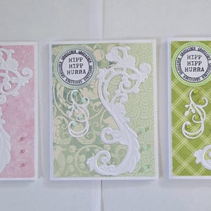 May include: Three decorative greeting cards with white filigree designs. Each card features a different background color: pink, light green, and green. The cards have a circular stamp with the text "HIPP HIPP HURRA".