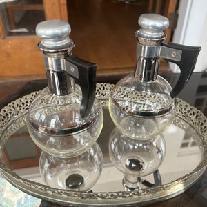 May include: Two clear glass bottles with silver tops and black handles. The bottles are sitting on a silver tray with a decorative design.
