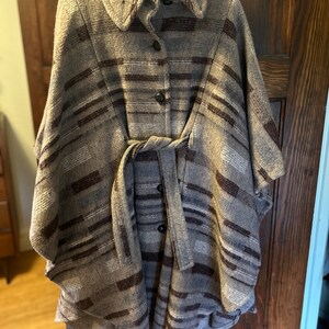May include: A brown and gray striped wool cape with a button closure and a tie belt. The cape has a collar and is lined with a brown fabric.