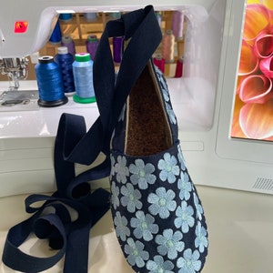 May include: A pair of blue canvas espadrilles with light blue embroidered flowers. The shoes have a navy blue ribbon that can be tied around the ankle.