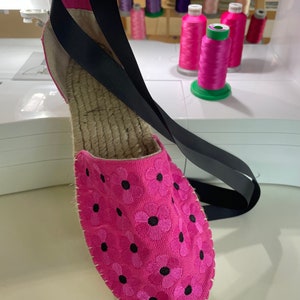 May include: Pink canvas espadrille shoe with black embroidered flowers. The shoe has a black strap that can be worn around the ankle.