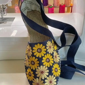 May include: A pair of blue espadrille shoes with yellow embroidered flowers. The shoes have a navy blue ribbon that ties around the ankle.