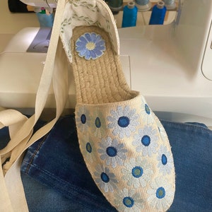 May include: A pair of beige espadrille shoes with blue and white embroidered flowers. The shoes have a white ribbon tied around the ankle.