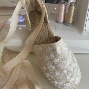 May include: A pair of beige espadrille shoes with white and yellow embroidered flowers. The shoes have a white ribbon that ties around the ankle.
