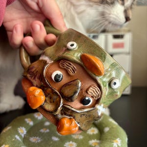 May include: A handmade ceramic mug featuring a sculpted face with textured eyebrows, a nose, and two eyes. The mug has a glossy green glaze, orange accents, and a handle. It is positioned above a round, green fabric coaster with a white daisy pattern.