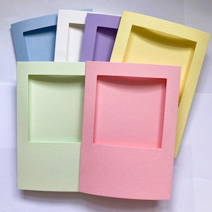 Op de afbeelding: Een verzameling van zes rechthoekige papieren frames in pastelkleuren, waaronder lichtblauw, wit, lavendel, geel, lichtgroen en roze. Elk frame heeft een getextureerd oppervlak en een grote centrale rechthoekige uitsparing voor foto's.