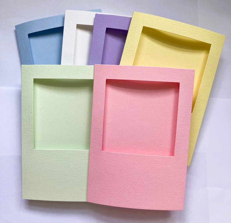 Op de afbeelding: Een verzameling van zes rechthoekige papieren frames in pastelkleuren, waaronder lichtblauw, wit, lavendel, geel, lichtgroen en roze. Elk frame heeft een getextureerd oppervlak en een grote centrale rechthoekige uitsparing voor foto's.