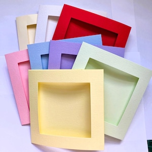 May include: A collection of square paper frames with a textured finish, arranged in a layered stack. The frames feature a variety of colours including red, white, cream, light blue, purple, light green, pink, and pale yellow, each with a central cutout.