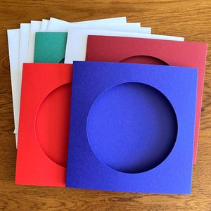 May include: A collection of coloured card mats with circular apertures. The mats are in red, blue, white, and green. They are arranged on a wooden surface.