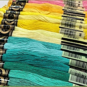 May include: A collection of colorful embroidery floss in shades of pink, yellow, and teal. Each skein is labeled with a black and gold band and a barcode. The floss is tightly wound and ready for crafting projects.