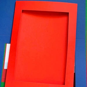 May include: A collection of coloured cardstock in red, blue, green, and white. A bright red rectangular frame sits atop the blue paper, creating a layered effect. Suitable for crafting and scrapbooking projects.