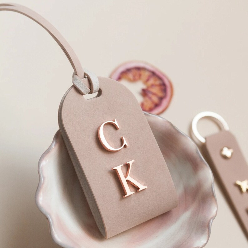 May include: A light brown leather luggage tag with a rose gold metal monogram "CK" on the front.