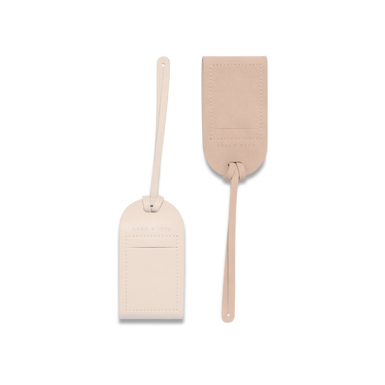May include: Two beige leather luggage tags with a stitched border and a small pocket. The tag on the left has the text "LANE & STYLE" embossed on it. The tag on the right has the text "WELL TRAVELED" embossed on it.