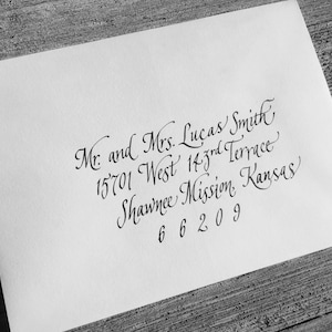 May include: A white envelope with elegant calligraphy. The text reads "Mr. and Mrs. Lucas Smith, 15701 West 143rd Terrace, Shawnee Mission, Kansas 66209". The envelope is placed on a wooden surface.