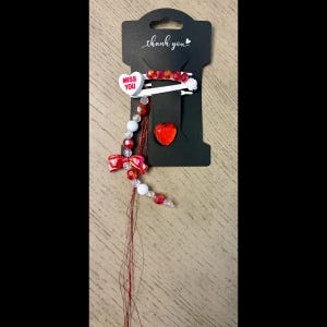May include: A black card with a white hair clip, a red heart-shaped gem, and a beaded strand. The hair clip has red and white beads and a heart-shaped charm that says "MISS YOU". The card says "thank you".