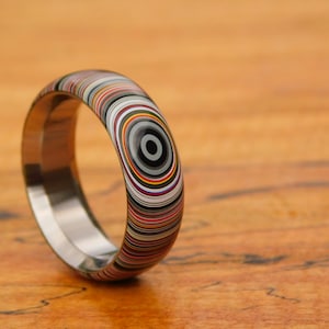 May include: A colorful ring with a concentric circle pattern in shades of black, white, red, yellow, and blue. The ring is made of titanium and has a polished finish.