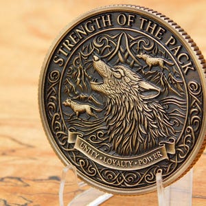 May include: A bronze coin featuring a detailed relief of a howling wolf and two smaller wolves. The coin's edge is ridged, and the text "STRENGTH OF THE PACK" and "UNITY LOYALTY POWER" are inscribed.