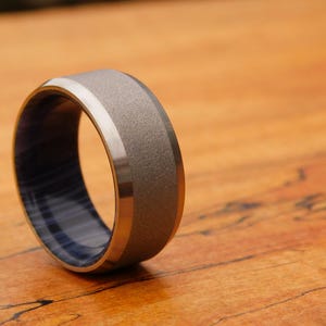 May include: A gray and silver ring with a blue and white wood inlay. The ring has a brushed finish and a beveled edge.