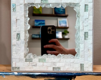 Könnte beinhalten: Quadratischer Spiegel mit einem Mosaikfliesenrahmen. Der Rahmen ist weiß mit kleinen rechteckigen Fliesen und transparenten Glasakzenten. Der Spiegel reflektiert eine Person, die ein Telefon hält.