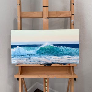 May include: A rectangular oil painting on canvas depicting a breaking ocean wave with turquoise and deep blue water, white sea foam, and a soft pale sky. The artwork is displayed on a light-coloured wooden artist easel with adjustable supports.