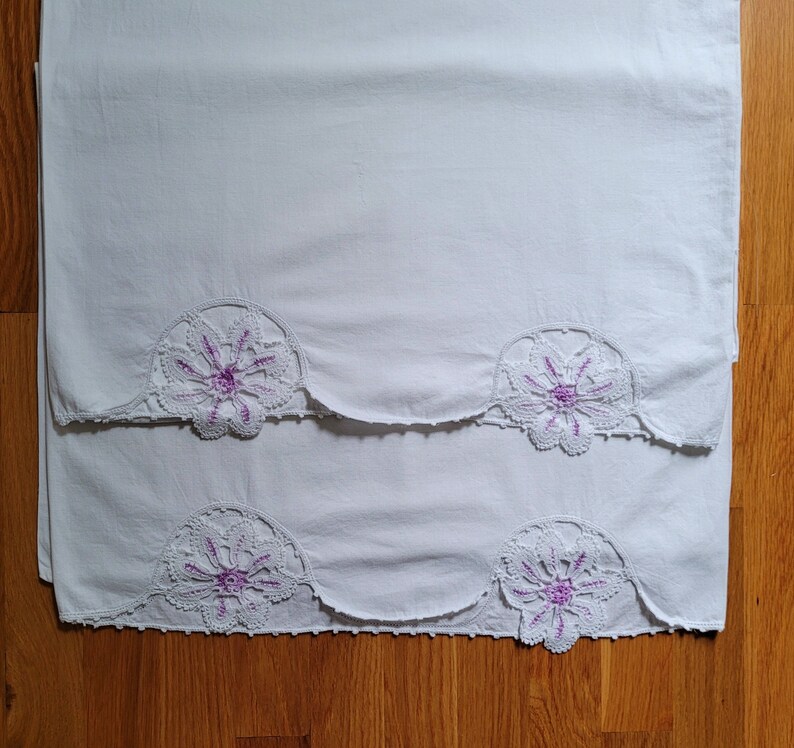 Puede incluir: Dos fundas de almohada de algod&oacute;n blanco con delicado encaje floral morado. El encaje tiene un patr&oacute;n de fest&oacute;n.
